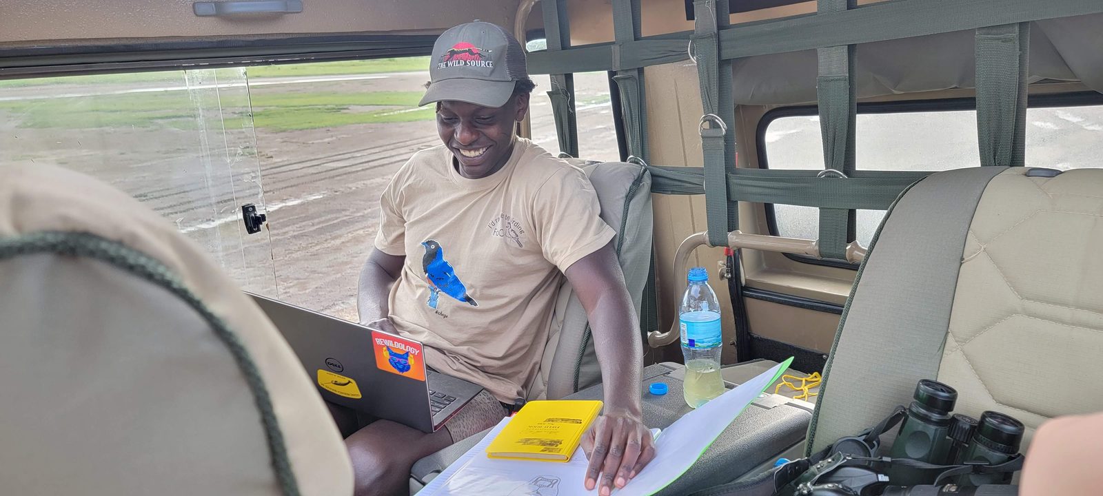 A guide in a safari vehicle recording sightings on a laptop and printed data sheets, binoculars beside him