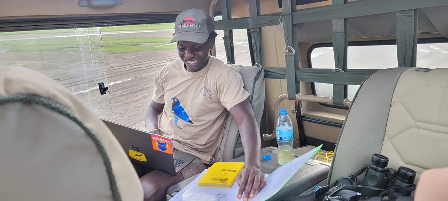 A guide in a safari vehicle recording sightings on a laptop and printed data sheets, binoculars beside him