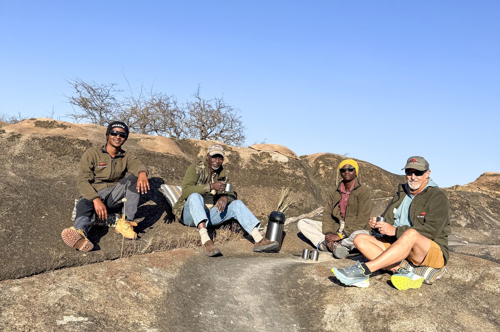 Four people sitting on a granite outcrop with a flask of coffee under a clear blue sky