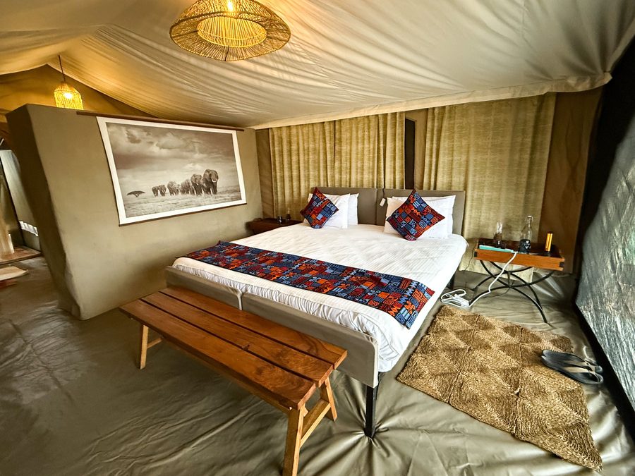 Interior of a guest tent: a made bed with patterned cushions, a wooden bench at its foot, a framed elephant print on the canvas wall