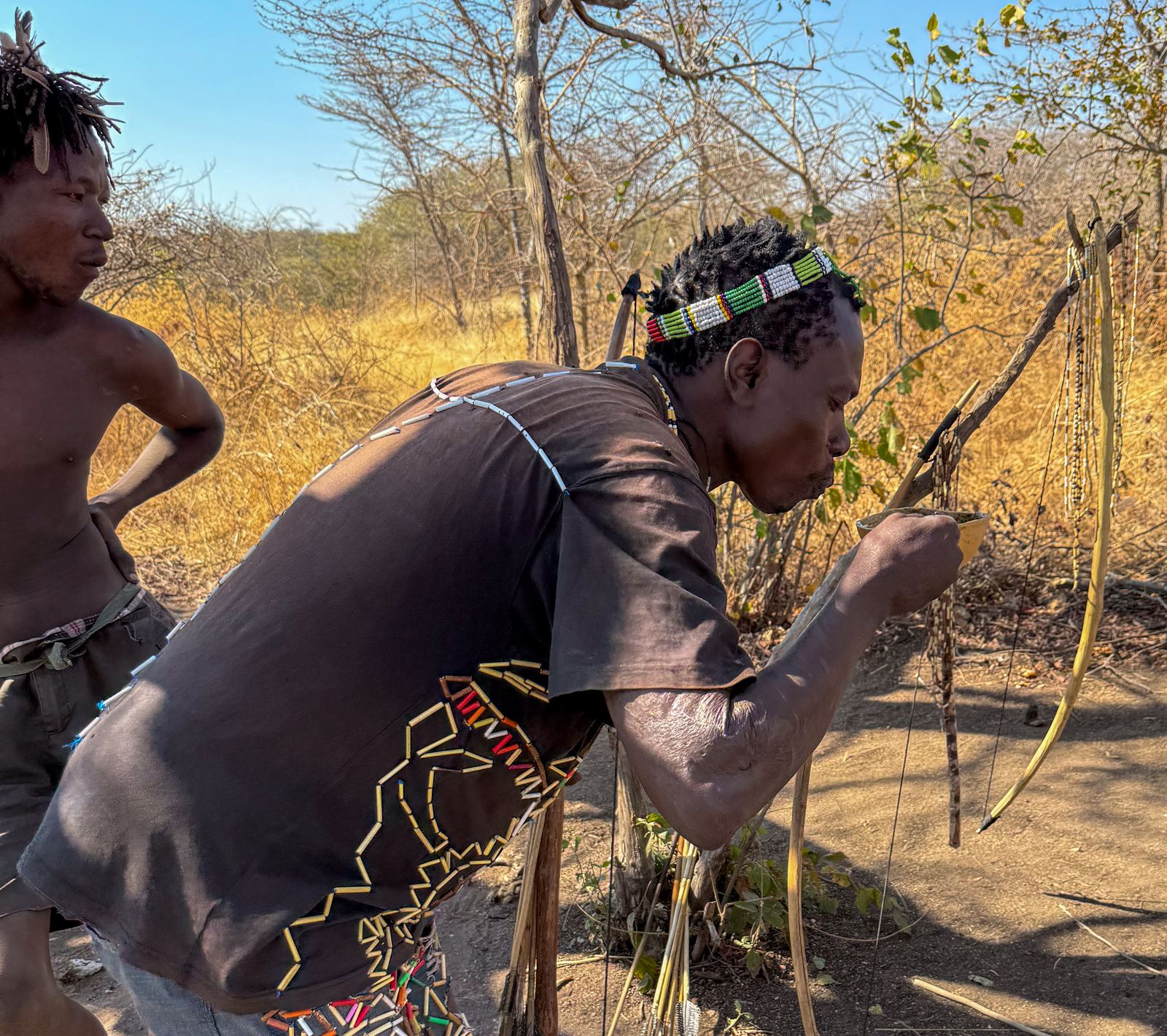A Hadzabe man in a beaded headband drawing a bow, with a second man in the background among dry bush