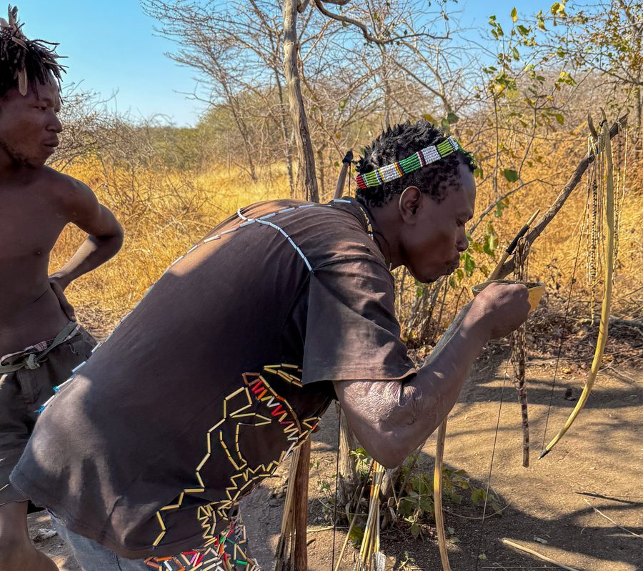 A Hadzabe man in a beaded headband drawing a bow, with a second man in the background among dry bush